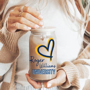 May include: A clear glass mason jar with a straw, filled with a brown liquid. The jar has a yellow heart with a blue outline and the text "Roger Williams University" in blue and yellow. The jar is being held by a person wearing a brown and white checked cardigan.