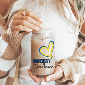 May include: A clear glass reusable iced coffee cup with a straw and a yellow heart with the words "University of Delaware" printed on it. The cup is being held by a person wearing a brown and white checkered cardigan.