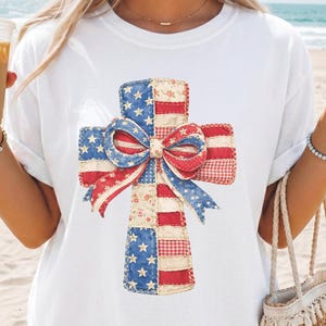 Könnte beinhalten: Ein weißes kurzärmeliges T-Shirt mit einer zentralen Grafik in Form eines Kreuzes, das aus rot-weiß-blauen Patchwork-Mustern mit Sternen und Streifen besteht und in der Mitte mit einer auffälligen Schleife verziert ist.