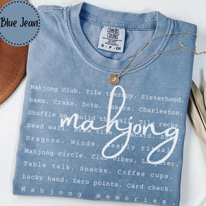 Op de afbeelding: Een blauw spijkerbroekkleurig t-shirt met het woord "mahjong" in een cursief lettertype. Het shirt heeft een lijst met mahjong-gerelateerde woorden in witte tekst. Een gouden ketting met een hanger ligt op het shirt.