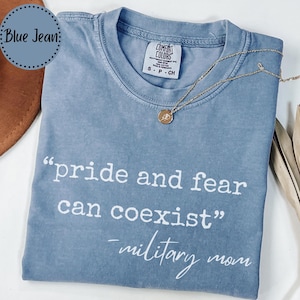Könnte beinhalten: Ein blaues Jeans-T-Shirt mit dem weißen Text "pride and fear can coexist - military mom". Eine goldene Halskette mit Anhänger liegt über dem Shirt. Das Shirt ist auf einer weißen Oberfläche gefaltet.