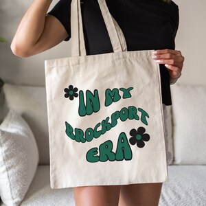 May include: A white canvas tote bag with a green and black design that says "*IN MY BROCKPORT ERA*" with a flower on either side of the text.