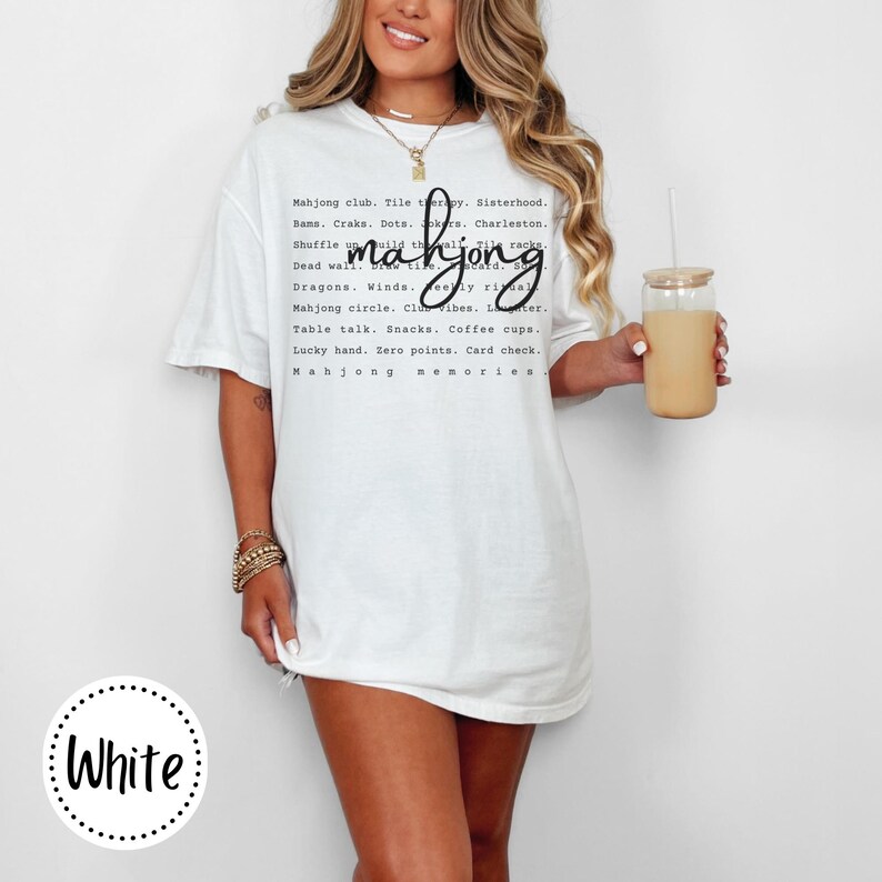 Op de afbeelding: Wit T-shirt met het woord "mahjong" in een cursief lettertype. Het shirt heeft een lijst met mahjong-gerelateerde woorden en zinnen in het zwart gedrukt. Een vrouw draagt het shirt en houdt een glas ijskoffie vast.
