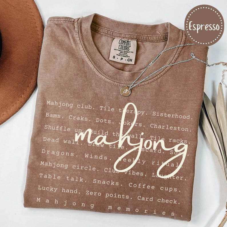 Op de afbeelding: Espresso-kleurige t-shirt met het woord "mahjong" in een cursief lettertype. De shirt heeft een lijst met mahjong-gerelateerde termen in witte tekst. Een zilveren ketting met een hanger is over het shirt gedrapeerd. De shirt is maat S.