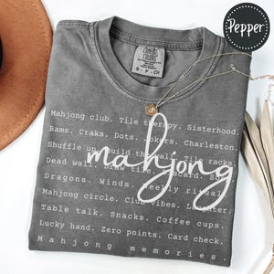 Op de afbeelding: Een grijs Comfort Colors T-shirt met het woord "mahjong" in een cursief lettertype. Het shirt heeft een lijst met mahjong-gerelateerde woorden in het wit, waaronder "Mahjong club", "Tile therapy" en "Lucky hand". Een gouden ketting met een hanger is over het shirt gedrapeerd.