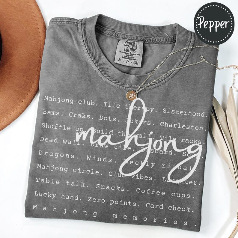 Op de afbeelding: Een grijs Comfort Colors T-shirt met het woord "mahjong" in een cursief lettertype. Het shirt heeft een lijst met mahjong-gerelateerde woorden in het wit, waaronder "Mahjong club", "Tile therapy" en "Lucky hand". Een gouden ketting met een hanger is over het shirt gedrapeerd.