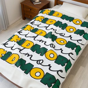 May include: White throw blanket with the word "Kidmore" repeated in a green and yellow pattern.