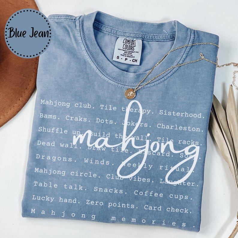 Op de afbeelding: Een blauw spijkerbroekkleurig t-shirt met het woord "mahjong" in een cursief lettertype. Het shirt heeft een lijst met mahjong-gerelateerde woorden in witte tekst. Een gouden ketting met een hanger ligt op het shirt.