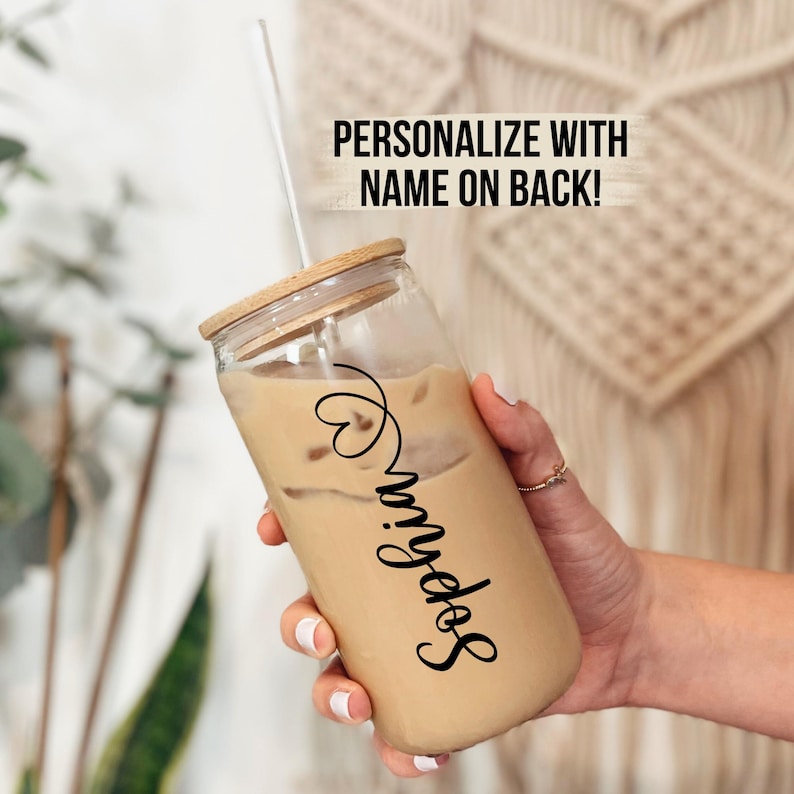 May include: A clear glass reusable iced coffee cup with a bamboo lid and a straw. The cup is filled with iced coffee and has the name "Sophia" written on the side in black cursive lettering. The text "PERSONALIZE WITH NAME ON BACK!" is written in black text on a white background.
