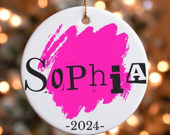 May include: White ceramic ornament with a gold string for hanging. The ornament features the name "Sophia" in black letters, with a bright pink brushstroke background. The year "-2024-" is printed below the name.