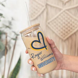 May include: A clear glass reusable iced coffee cup with a bamboo lid and a straw. The cup has a yellow heart with a blue outline and the text "Roger Williams University" in blue and yellow lettering.