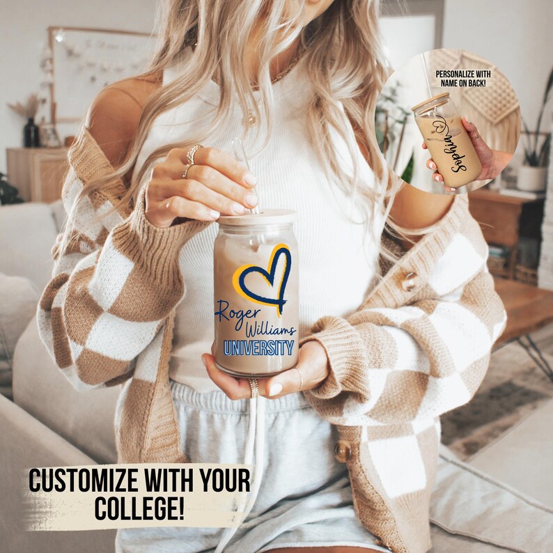 May include: A clear glass mason jar with a straw, filled with a brown liquid. The jar has a yellow heart with a blue outline and the text "Roger Williams University" in blue and yellow. The jar is being held by a person wearing a brown and white checked cardigan.