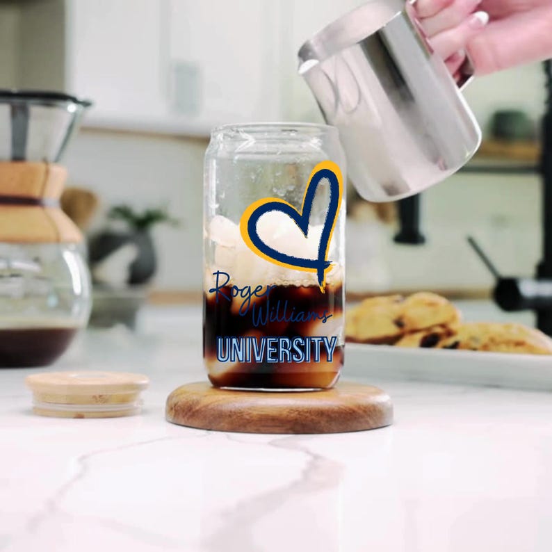 May include: A clear glass with a blue and yellow heart design and the text "Roger Williams University" is filled with ice and coffee. Cream is being poured into the glass from a silver jug.