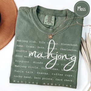 Op de afbeelding: Mosgroen t-shirt met witte tekst over Mahjong. Het shirt heeft een lijst met Mahjong-termen en -zinnen, waaronder "Mahjong club", "Shuffle up" en "Lucky hand". Een gouden ketting met een hanger is over het shirt gedrapeerd.