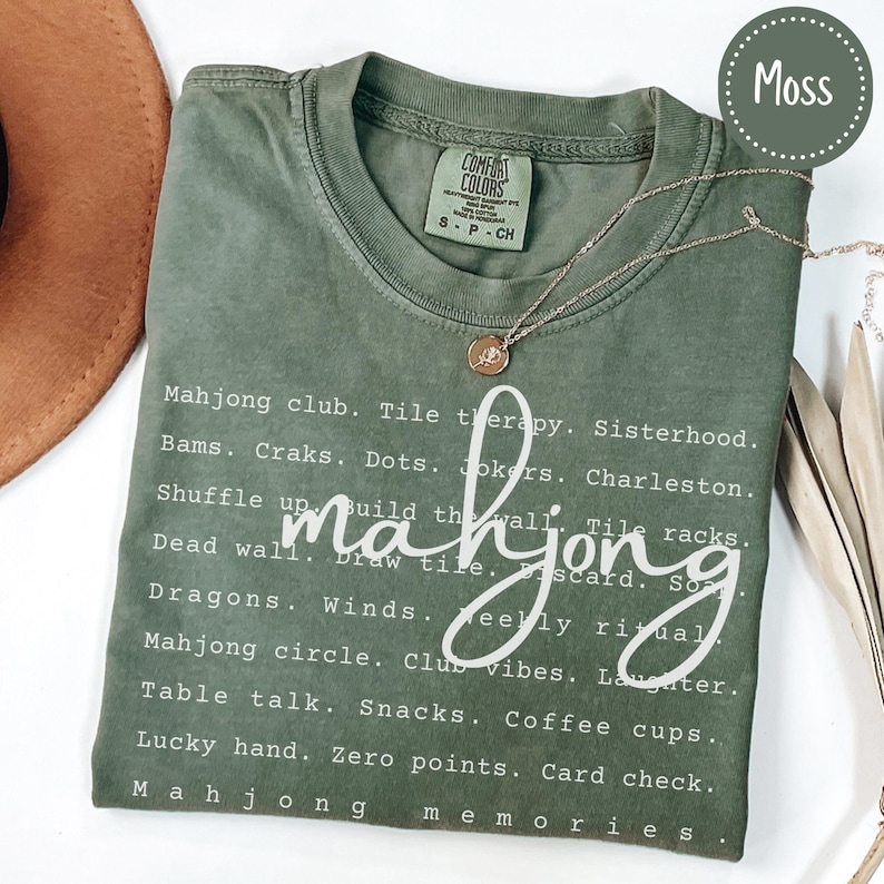 Op de afbeelding: Mosgroen t-shirt met witte tekst over Mahjong. Het shirt heeft een lijst met Mahjong-termen en -zinnen, waaronder "Mahjong club", "Shuffle up" en "Lucky hand". Een gouden ketting met een hanger is over het shirt gedrapeerd.
