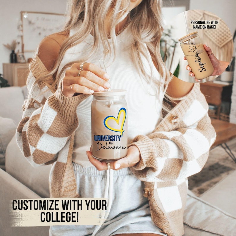 May include: A clear glass reusable iced coffee cup with a straw and a yellow heart with the words "University of Delaware" printed on it. The cup is being held by a person wearing a brown and white checkered cardigan.
