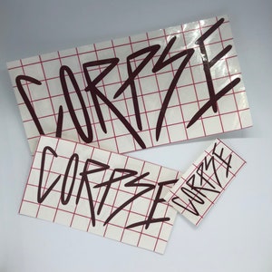 Op de afbeelding: Drie witte stickers met het woord "CORPSE" geschreven in zwarte stift op een rode geruite achtergrond.