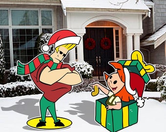 May include: A Christmas-themed yard decoration featuring a cartoon character wearing a red and green outfit and a Santa hat, standing next to a large green gift box with a cartoon character peeking out of the top.