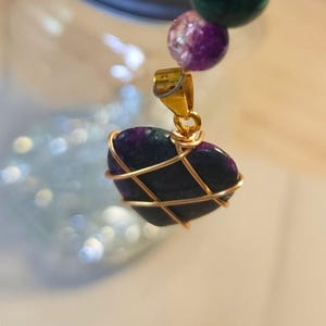 May include: A beaded bracelet with a heart-shaped pendant. The bracelet features green, purple, and clear beads. The heart-shaped pendant is a dark purple stone wrapped in gold wire.