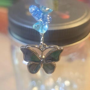 May include: A decorative jar with a silver butterfly charm and blue beads. The butterfly charm has a silver outline with blue and green accents. The beads are various shades of blue. The jar has a black lid and is filled with small, clear objects.