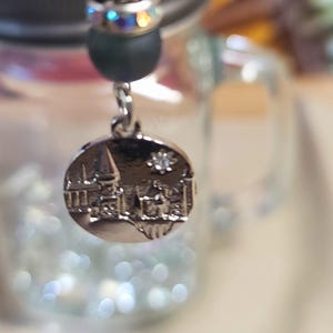 May include: A silver charm with a detailed castle design hangs from a black bead. The charm is suspended from a dark green bead and a bead with iridescent accents. The charm is attached to a black bead.
