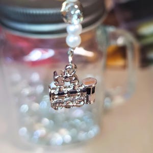 May include: A silver train charm hangs from a beaded strand of white pearls and faceted crystals. The charm is suspended in front of a clear glass jar with a silver lid, filled with small, sparkling beads. The background is blurred with colorful accents.
