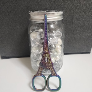 May include: Iridescent Eiffel Tower-shaped scissors with a silver jar filled with clear and pearl-colored beads in the background. The scissors have a decorative design and are made of metal. The jar has a silver lid.