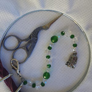 May include: A silver embroidery hoop with a white fabric background. A pair of silver stork scissors and a beaded charm with green and clear beads are inside the hoop. A maroon thread is visible.