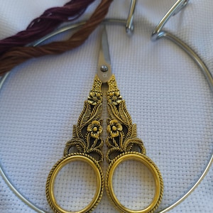 May include: Close-up of gold-colored embroidery scissors with floral detailing, resting on white cross-stitch fabric within a metal hoop. Strands of brown and burgundy embroidery floss are visible, suggesting a crafting project.