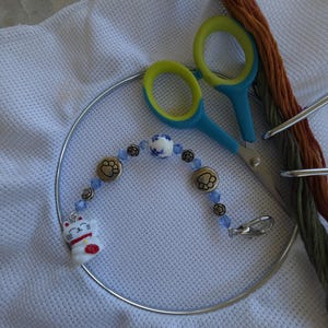 May include: A beaded needle minder featuring a white ceramic lucky cat charm, blue crystal beads, silver-toned paw print charms, and a floral bead. The accessory includes a metal lobster clasp and is displayed with embroidery scissors and thread.