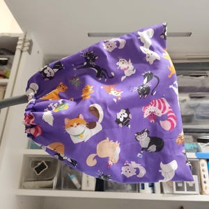 May include: A purple fabric item with a repeating pattern of various cartoon cats in different poses and colors. The cats include orange, black, white, and striped designs. The item is gathered at one end.