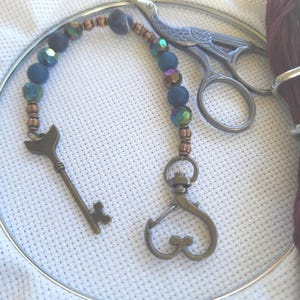 May include: A beaded scissor fob with a key charm and a lobster clasp, alongside a pair of embroidery scissors. The fob features dark blue and iridescent beads with bronze-colored accents. The key charm is a bronze color.