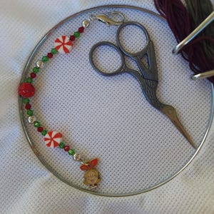 May include: A metal embroidery hoop containing a pair of stork-shaped scissors, a bundle of dark red and green embroidery floss, and a beaded charm strand featuring red and white peppermint beads, green crystals, and a small cartoon girl charm.