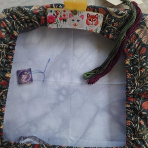 May include: A square embroidery frame cover featuring a dark teal fabric with a floral pattern, secured by a yellow plastic clip decorated with cat faces and flowers. A small purple needle minder and two strands of embroidery floss are attached.