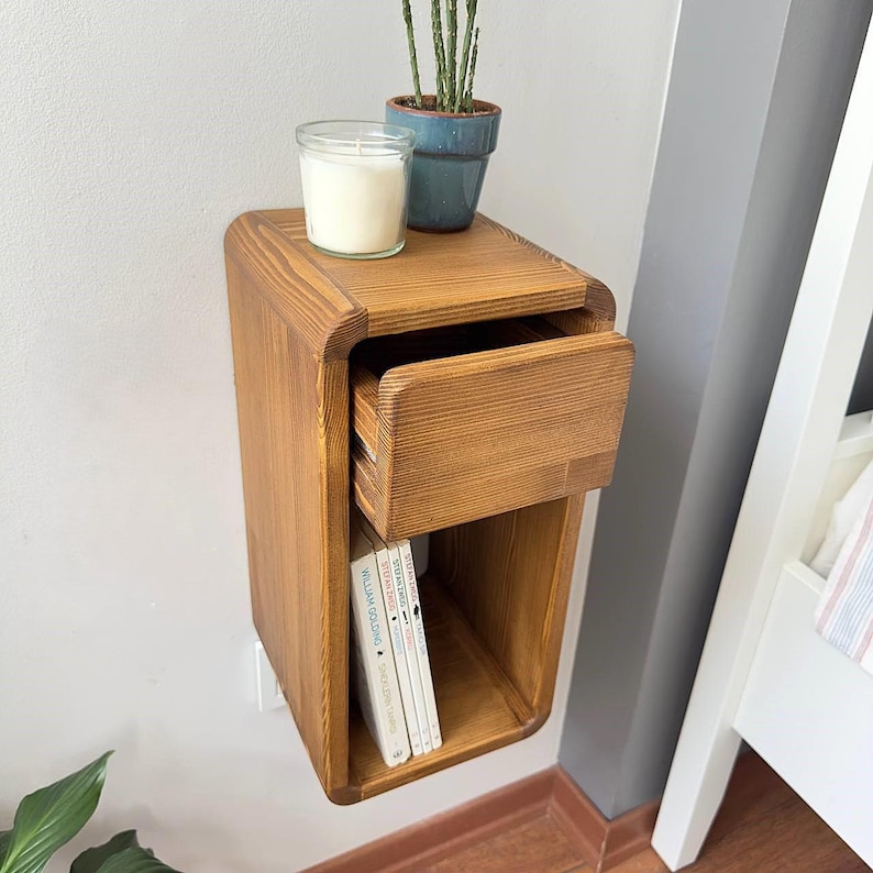 Puede incluir: Una mesita de noche de madera montada en la pared con un caj&oacute;n y un estante abierto. La mesita de noche es de madera marr&oacute;n claro y tiene bordes redondeados. Una peque&ntilde;a maceta azul con una planta y una vela est&aacute;n encima. Se ven libros.