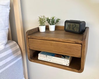 May include: A floating wooden nightstand with a drawer and open shelf. The nightstand is stained a warm brown and features rounded edges. Two small potted plants and a digital clock sit on top. Books are visible on the shelf below.
