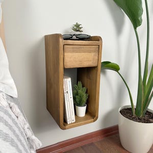 May include: A wooden wall-mounted nightstand with a drawer and a shelf. The nightstand is stained a dark brown color and has a curved front edge. The shelf is holding a stack of books and a small potted plant. A pair of sunglasses is sitting on top of the drawer.