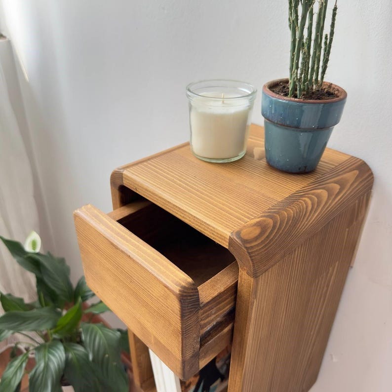 Puede incluir: Una mesita de noche de madera clara con un caj&oacute;n abierto. La mesita tiene bordes redondeados. Una vela encendida en un tarro de cristal y una peque&ntilde;a planta en maceta est&aacute;n encima.