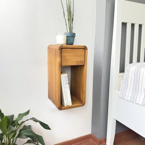 Può includere: Un comodino in legno a parete con un cassetto e uno scaffale. Il comodino è in legno marrone chiaro e ha un design curvo. C'è una piccola pianta in vaso, una candela e una pila di libri sullo scaffale.