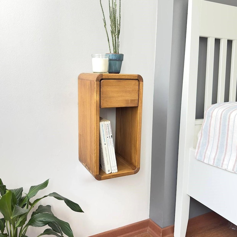 Puede incluir: Una mesita de noche flotante de madera con un caj&oacute;n y una estanter&iacute;a abierta. La mesita de noche es de madera marr&oacute;n claro y est&aacute; montada en una pared blanca. Una peque&ntilde;a planta en maceta y una vela est&aacute;n encima, y hay libros en el estante.