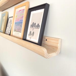Puede incluir: Una estantería de madera con un borde curvo, montada en una pared blanca. La estantería contiene varias imágenes enmarcadas, incluyendo una con un marco negro y una impresión de una pila de piedras.
