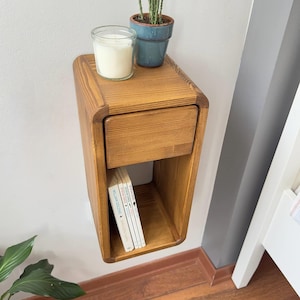 Puede incluir: Una mesita de noche flotante de madera con un caj&oacute;n y una estanter&iacute;a abierta. La mesita de noche es de madera marr&oacute;n claro y tiene bordes redondeados. Una vela en un tarro de cristal y una peque&ntilde;a planta en maceta est&aacute;n encima. Se ven libros en el estante.