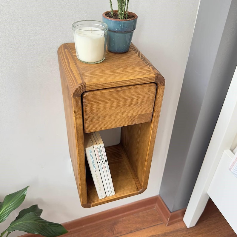 Puede incluir: Una mesita de noche flotante de madera con un caj&oacute;n y una estanter&iacute;a abierta. La mesita de noche es de madera marr&oacute;n claro y tiene bordes redondeados. Una vela en un tarro de cristal y una peque&ntilde;a planta en maceta est&aacute;n encima. Se ven libros en el estante.
