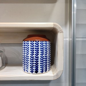 May include: A white ceramic jar with a blue and white leaf pattern and a brown top sits on a wooden shelf.