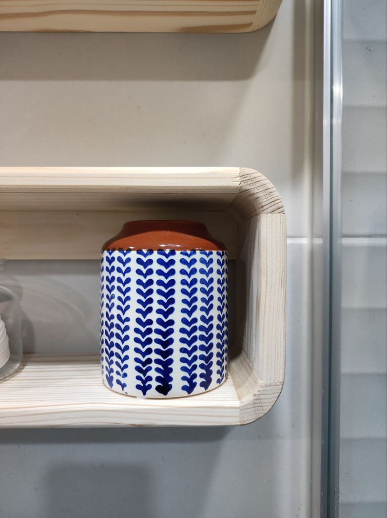May include: A white ceramic jar with a blue and white leaf pattern and a brown top sits on a wooden shelf.