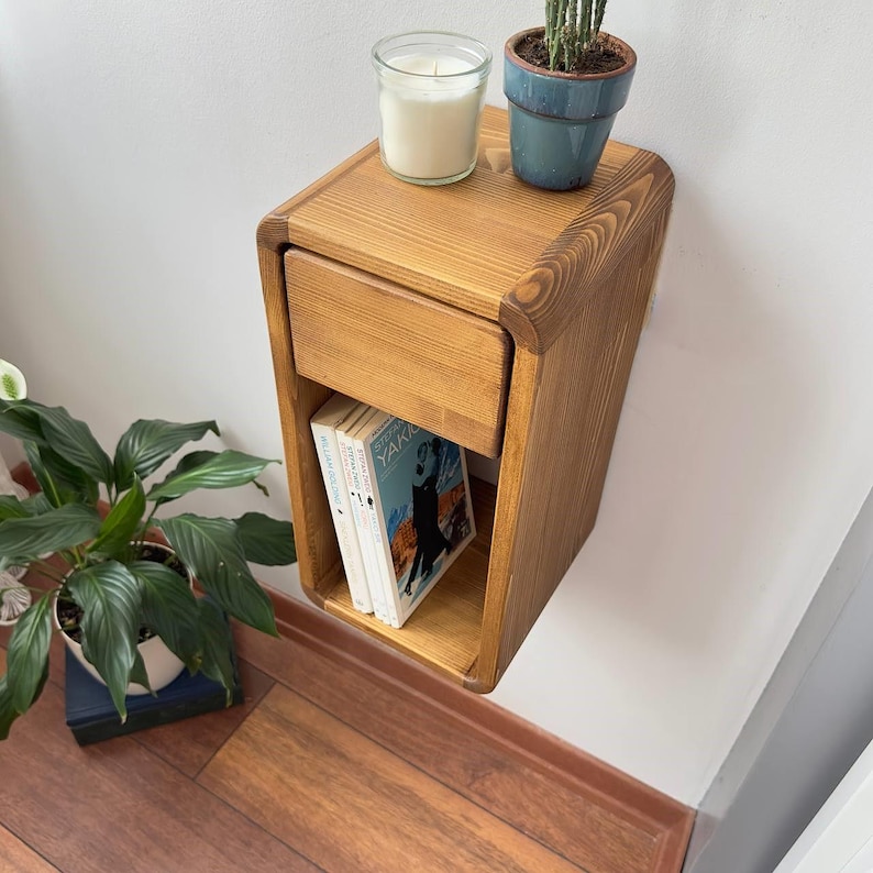 Puede incluir: Una mesita de noche flotante de madera con un caj&oacute;n y una estanter&iacute;a abierta. Una vela en un tarro de cristal y una peque&ntilde;a planta en maceta est&aacute;n encima. Dos libros son visibles en la estanter&iacute;a. La mesita est&aacute; montada en una pared blanca.