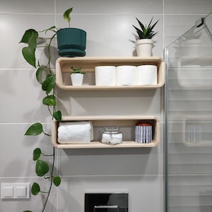 May include: Two wooden shelves with rounded edges, mounted on a white tiled wall. The top shelf holds three rolls of toilet paper and a small green potted plant. The bottom shelf has a white towel, a glass jar with cotton balls, and a blue and white patterned jar.