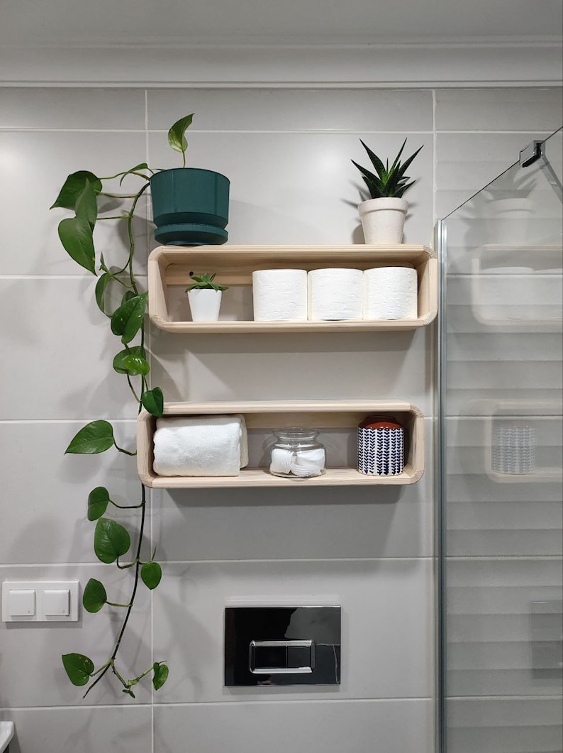 May include: Two wooden shelves with rounded edges, mounted on a white tiled wall. The top shelf holds three rolls of toilet paper and a small green potted plant. The bottom shelf has a white towel, a glass jar with cotton balls, and a blue and white patterned jar.