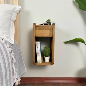 Puede incluir: Una mesita de noche de madera montada en la pared con un cajón y un estante. El estante tiene una pequeña planta en maceta y una pila de libros. La mesita de noche está hecha de madera natural con un acabado marrón claro.