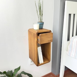Puede incluir: Una mesita de noche flotante de madera con un caj&oacute;n y una estanter&iacute;a abierta. La mesita de noche es de madera marr&oacute;n claro y est&aacute; montada en una pared gris. Una peque&ntilde;a planta en maceta, una vela y libros est&aacute;n encima.